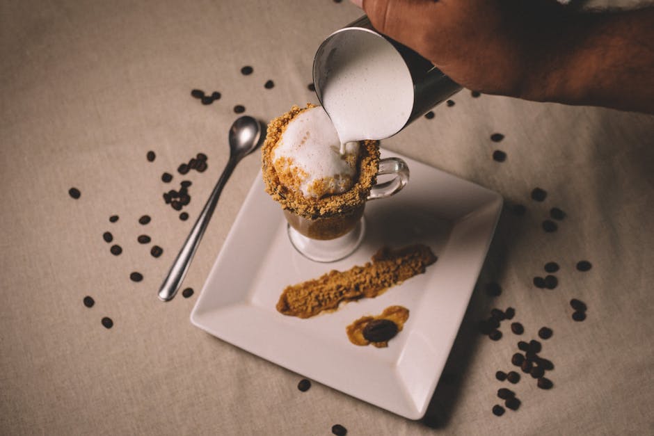 Close-up of creamy coffee in a glass cup with frothy milk being poured, surrounded by coffee beans.