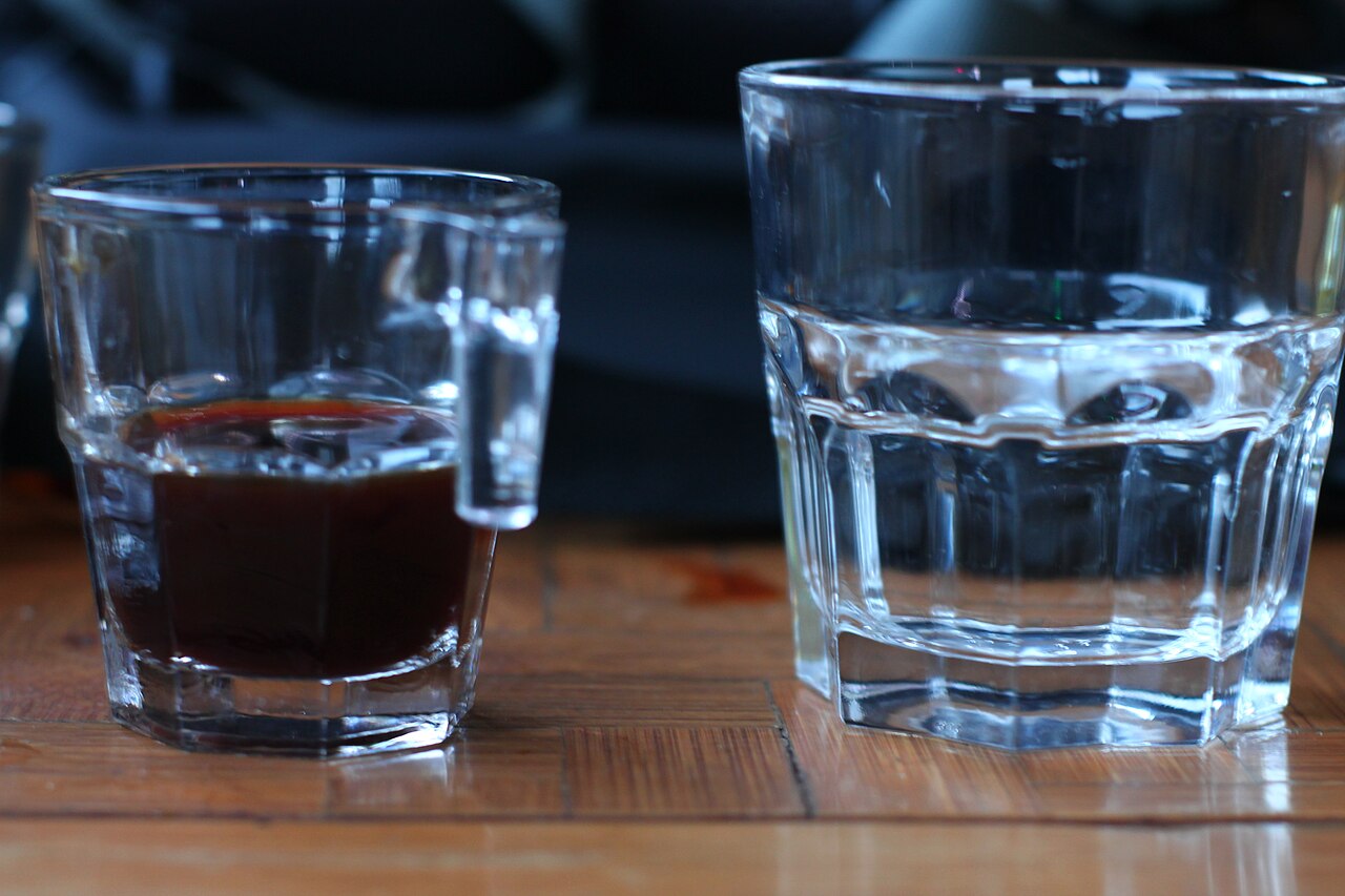Two glasses side by side — one of cold brew (darker, smoother surface) and one of iced coffee (slightly redder, more lively surface) — with labels