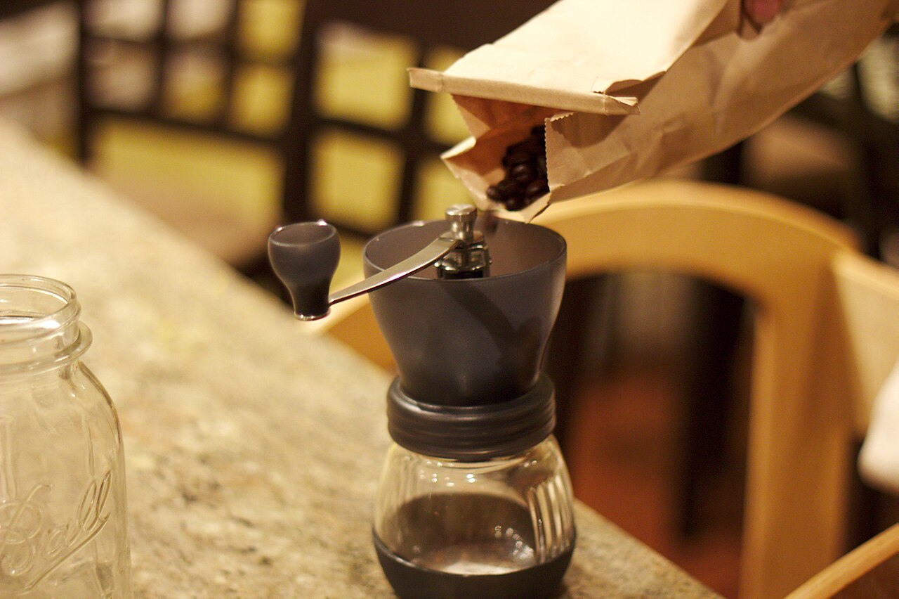 Whole coffee beans poured from a paper bag into a hand-crank burr grinder, with an empty glass mason jar ready on the counter for cold brew steeping