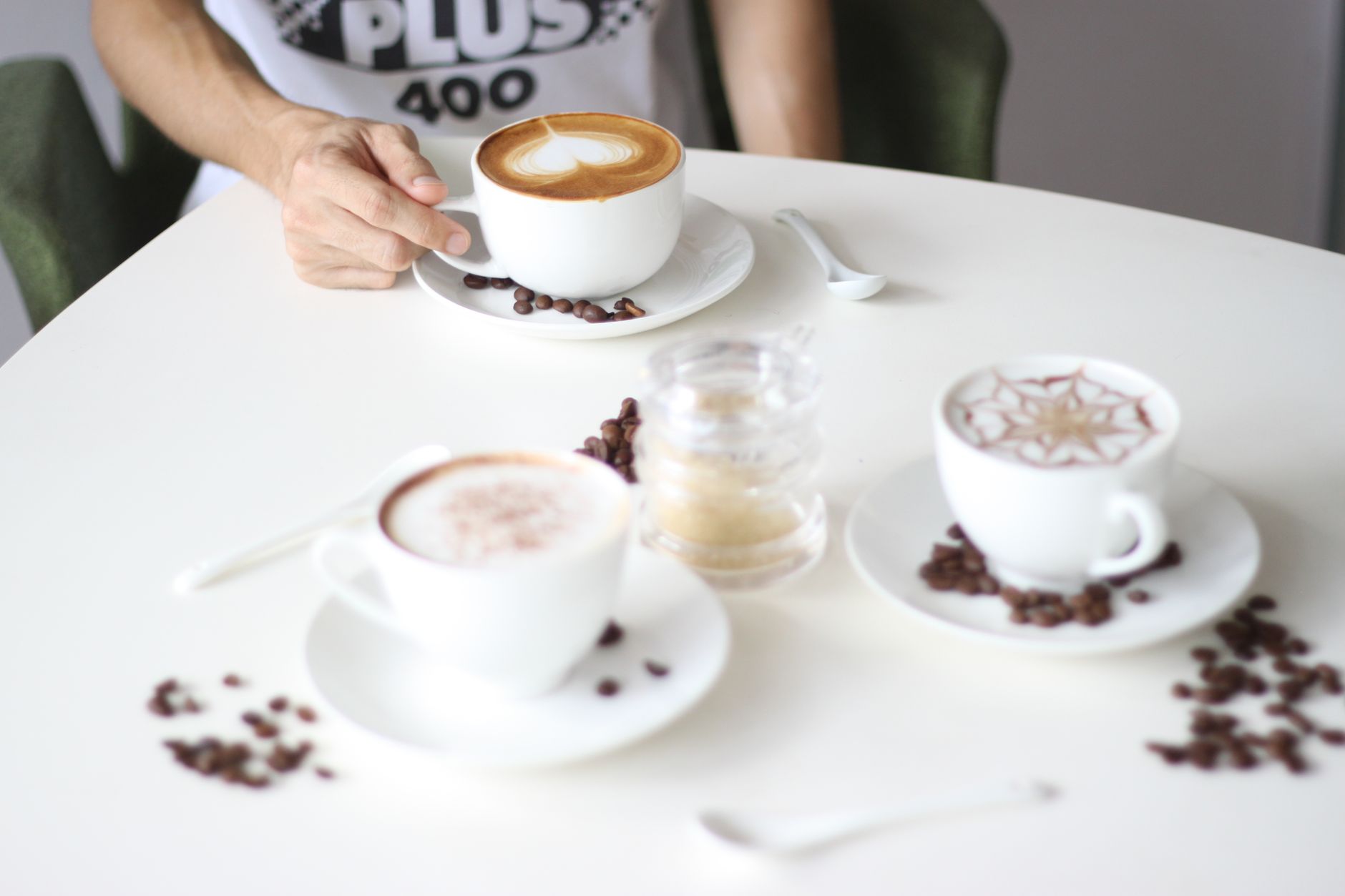 Three cappuccinos with decorative latte art on a cafe table, with roasted coffee beans scattered around the saucers.