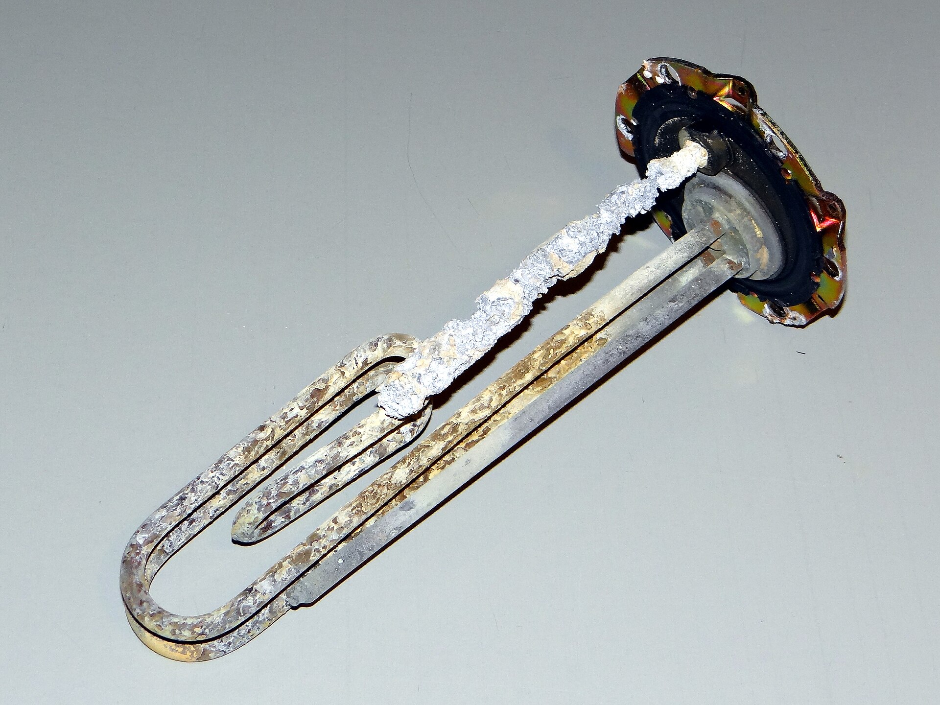 An electric heating element pulled from a boiler, thickly encrusted with limescale beside a corroded magnesium anode - the calcium and magnesium scale that untreated water deposits inside espresso machine boilers