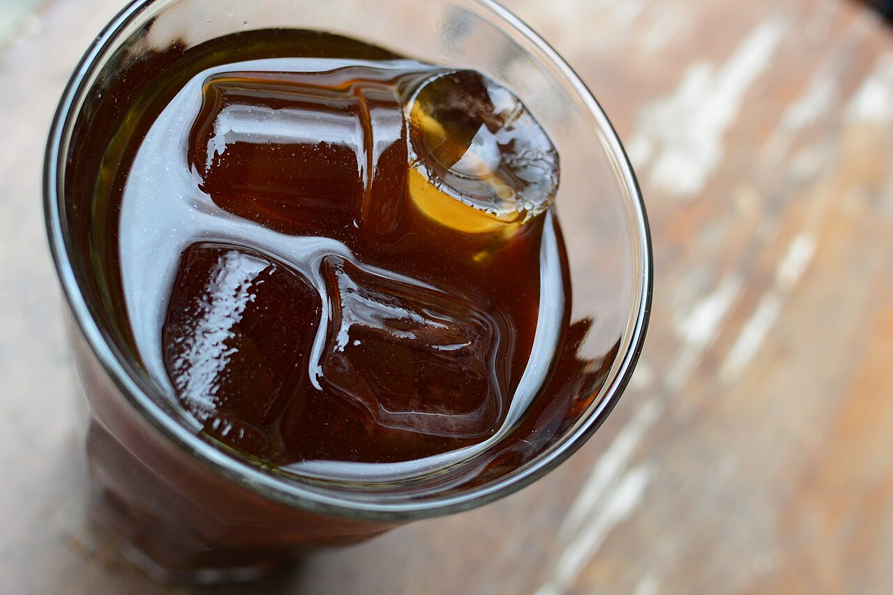 A tall glass of cold brew coffee over ice cubes, seen from above on a weathered wooden surface