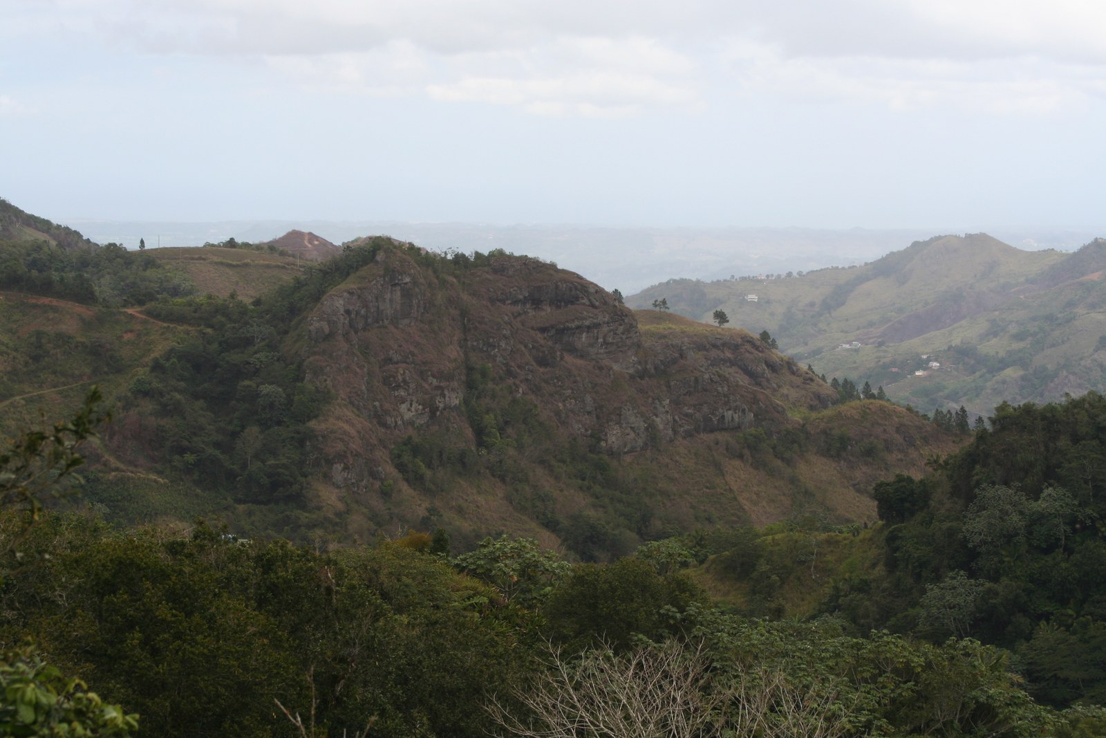 A rocky hill with brown, dry slopes at Collores, Juana Díaz, with scattered houses on the ridges beyond and hazy lowland on the horizon.