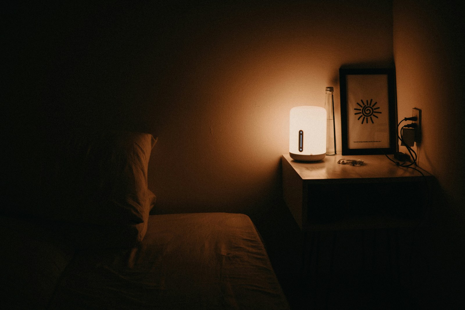 A dark room with a bed and one lit lamp on a bedside table.