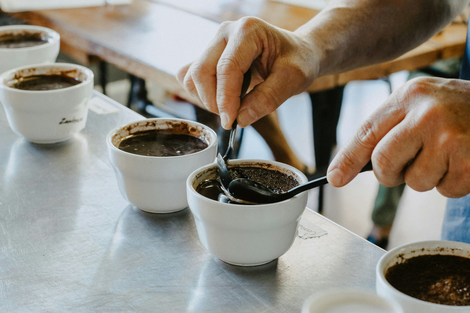 Two hands holding spoons over white cupping bowls, one spoon breaking the crust of wet grounds, with further bowls on the steel table around them.
