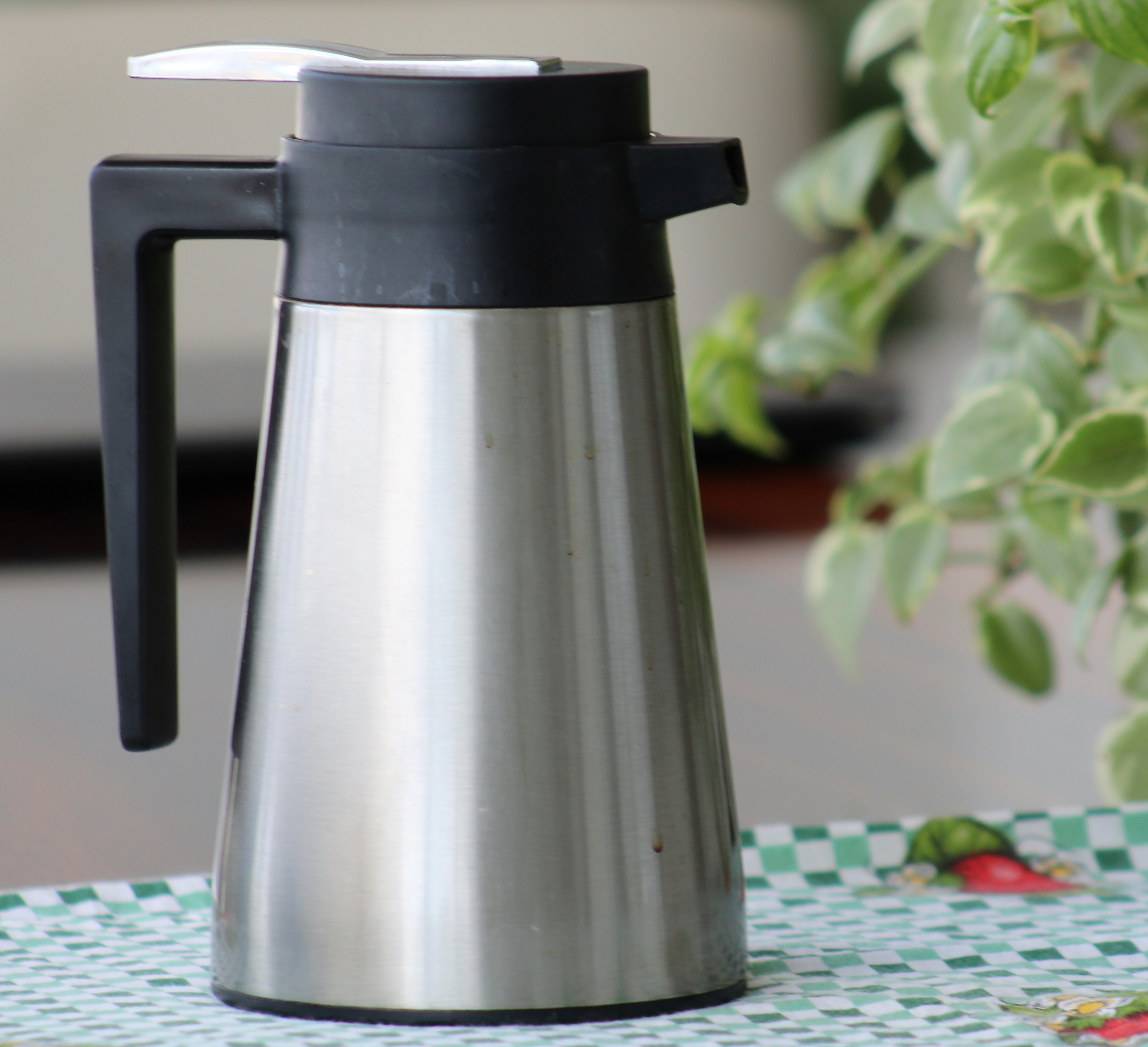 A stainless steel thermal coffee carafe with a black lid and handle, standing on a checked tablecloth.
