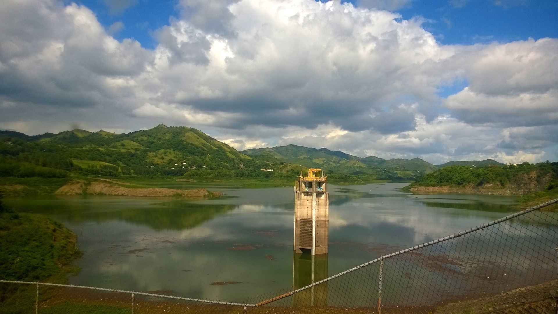 The concrete intake tower of Lago Toa Vaca in Villalba, seen through a chain-link fence, with green hills behind under cumulus cloud.