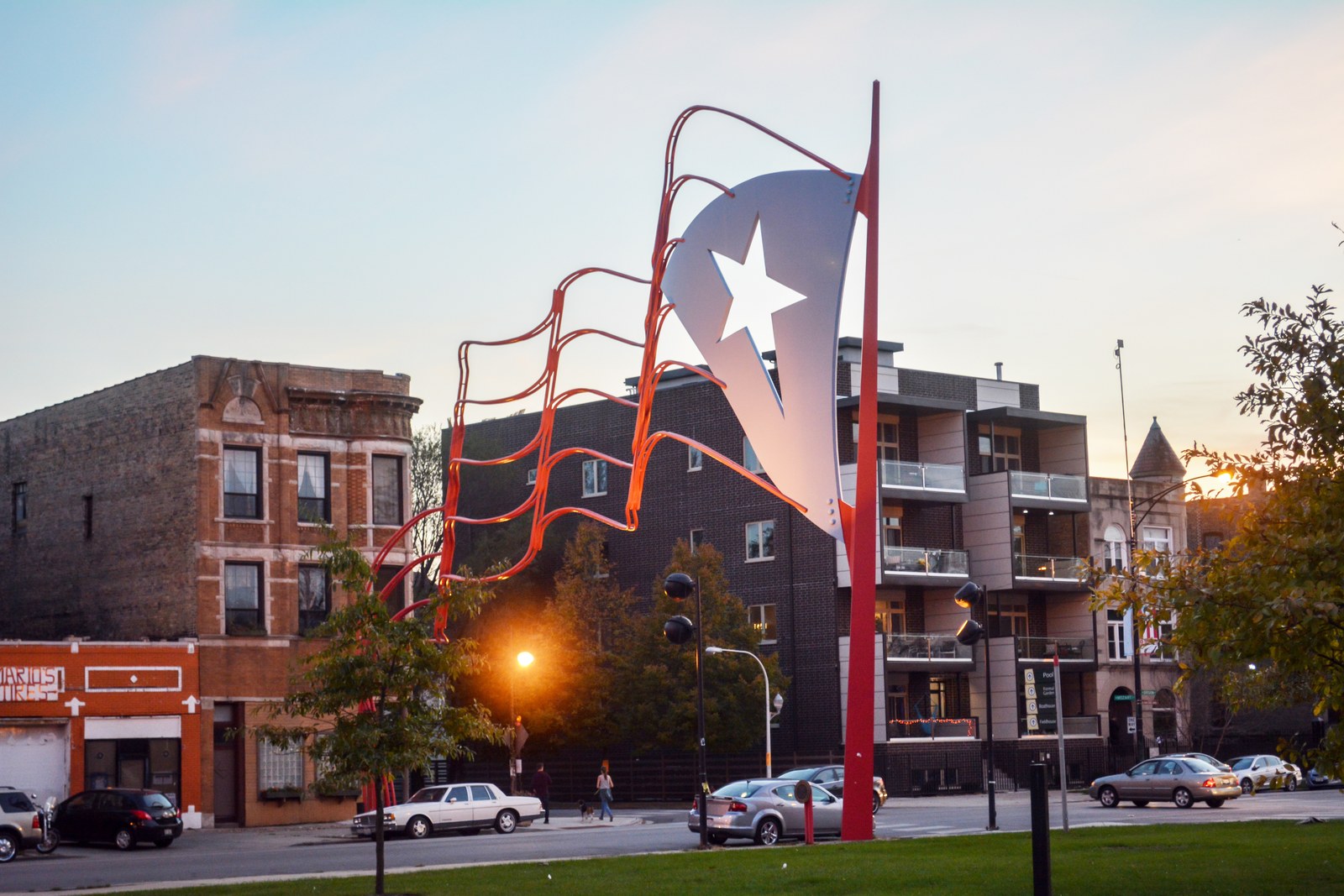 A giant steel Puerto Rican flag sculpture on Paseo Boricua in Chicago at dusk, with brick apartment buildings and parked cars behind.