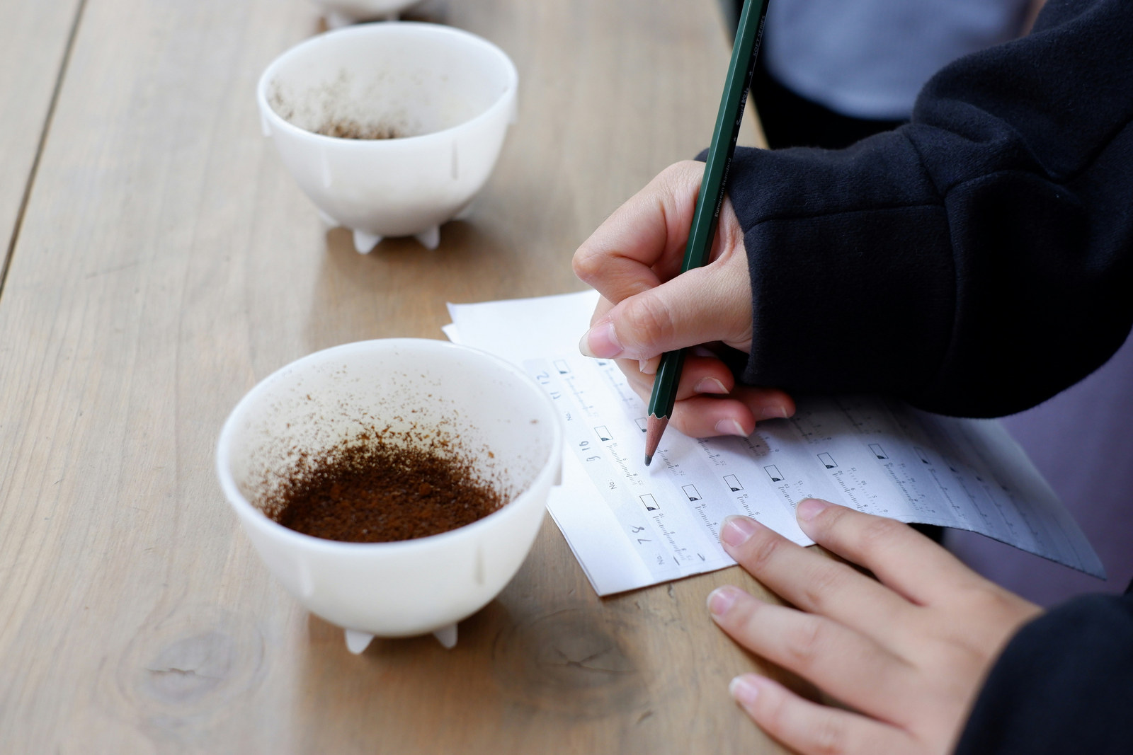 A hand recording scores with a pencil on a printed tasting form with numbered intensity scales, beside two cupping bowls of ground coffee.