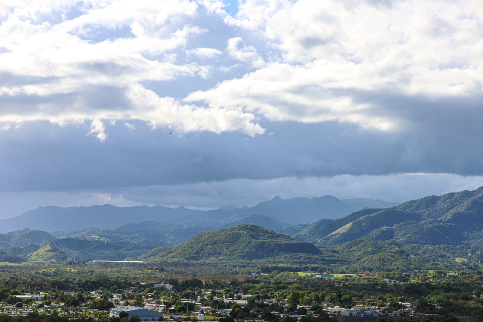 The southern face of the Cordillera Central under rain cloud, photographed from Juana Díaz, not Villalba, with green foothills and a town in the foreground.