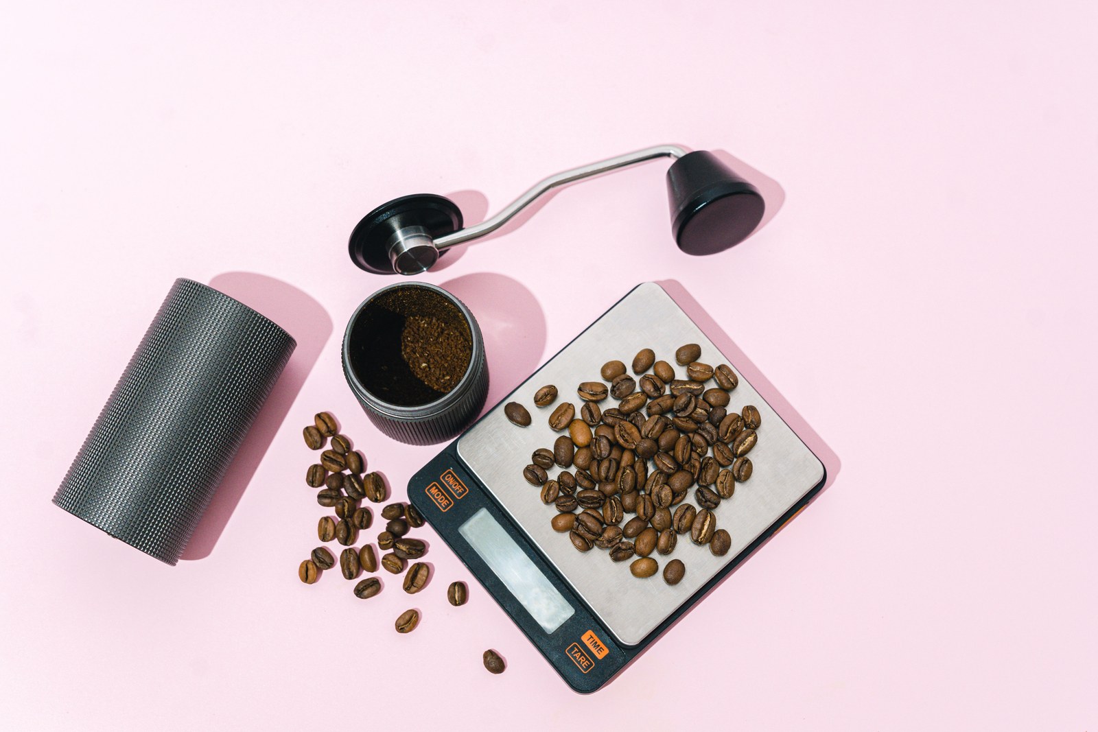 A hand coffee grinder taken apart, its catch cup of fresh grounds, and whole beans on a stainless-topped digital scale with tare and time buttons and a blank display.