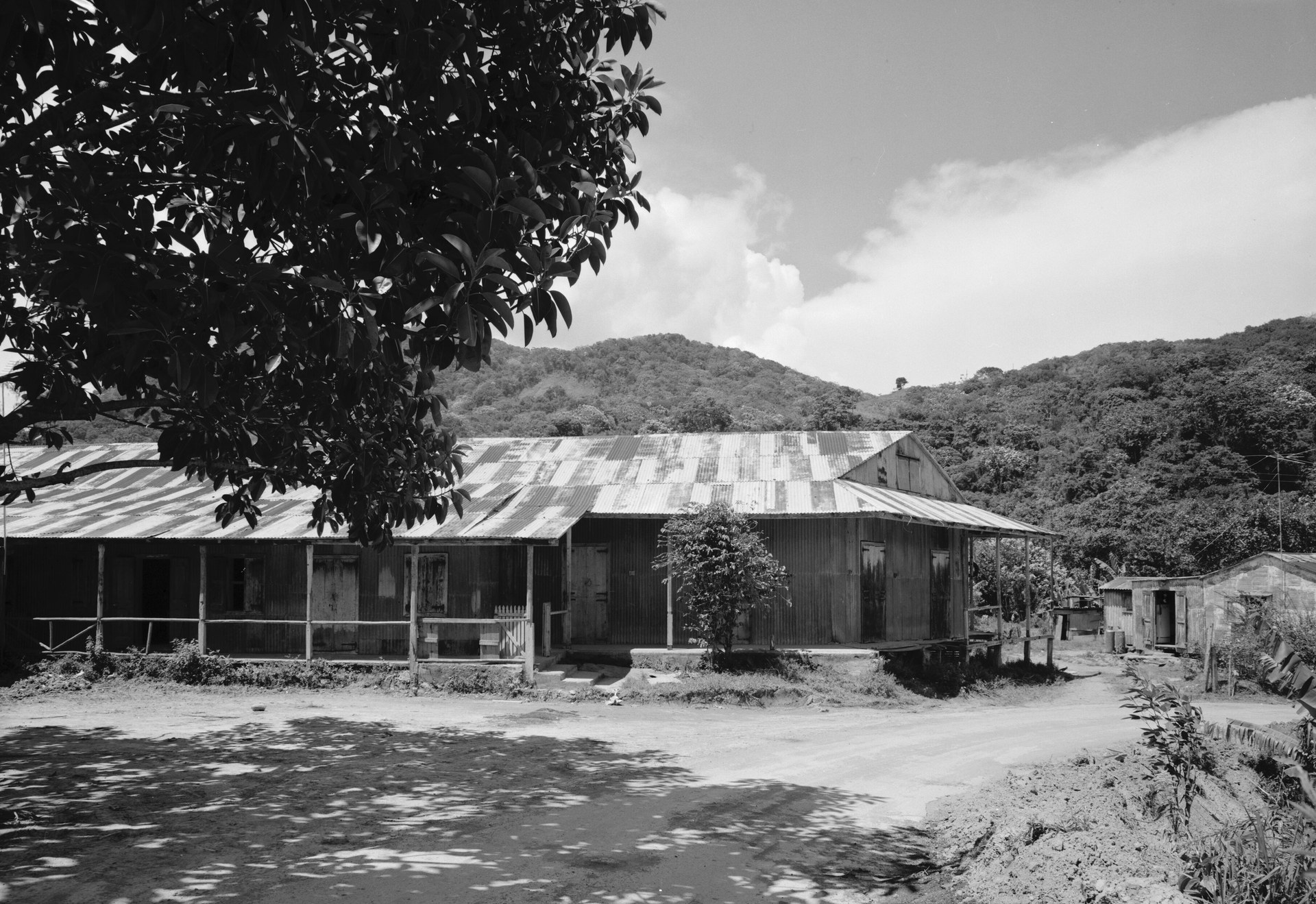 A long coffee mill with a corrugated-iron roof and a timber porch, beside a dirt road below wooded hills.