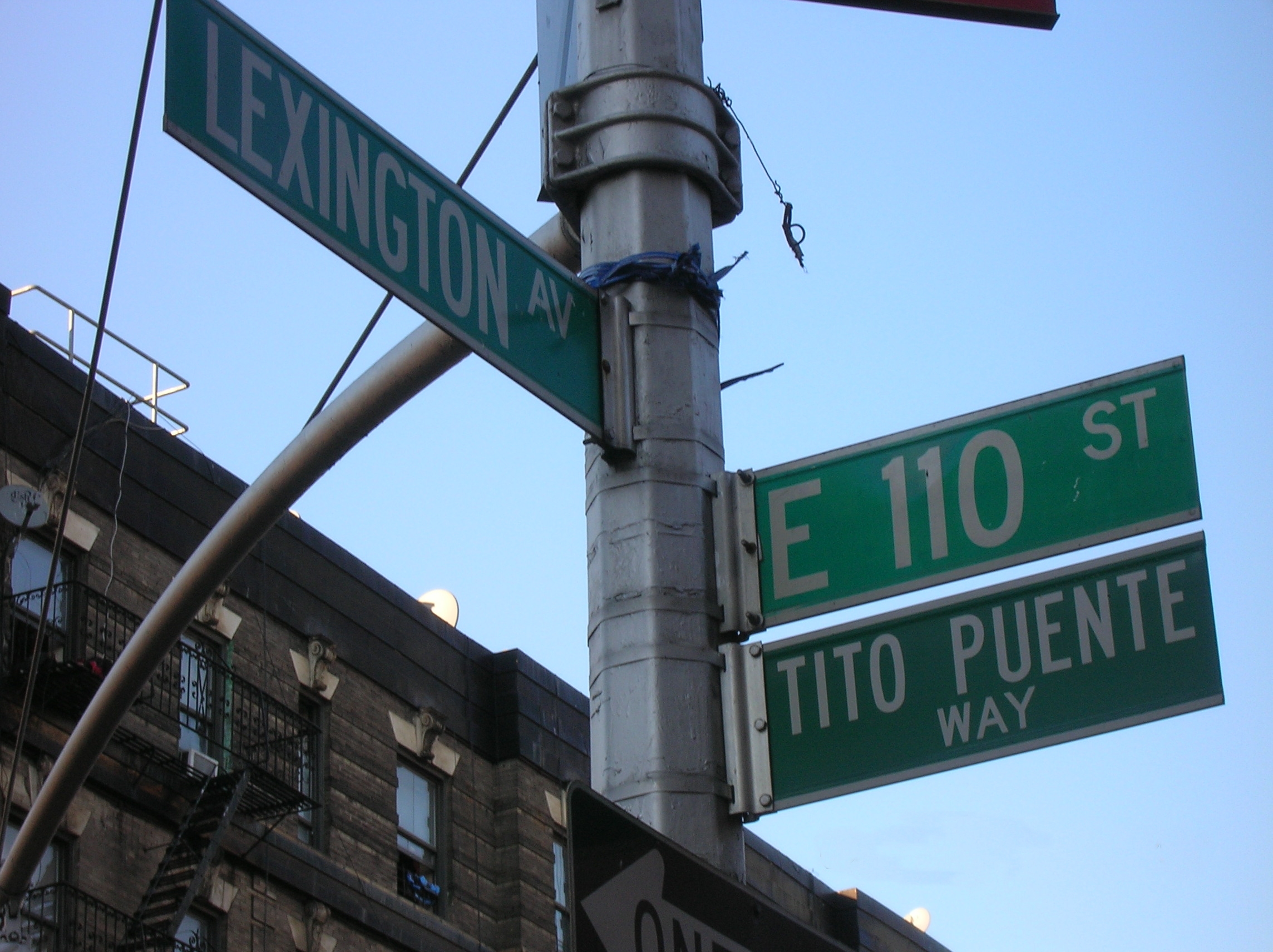 The East 110th Street and Tito Puente Way street signs at Lexington Avenue in East Harlem, New York, with a brick tenement and its fire escapes behind.
