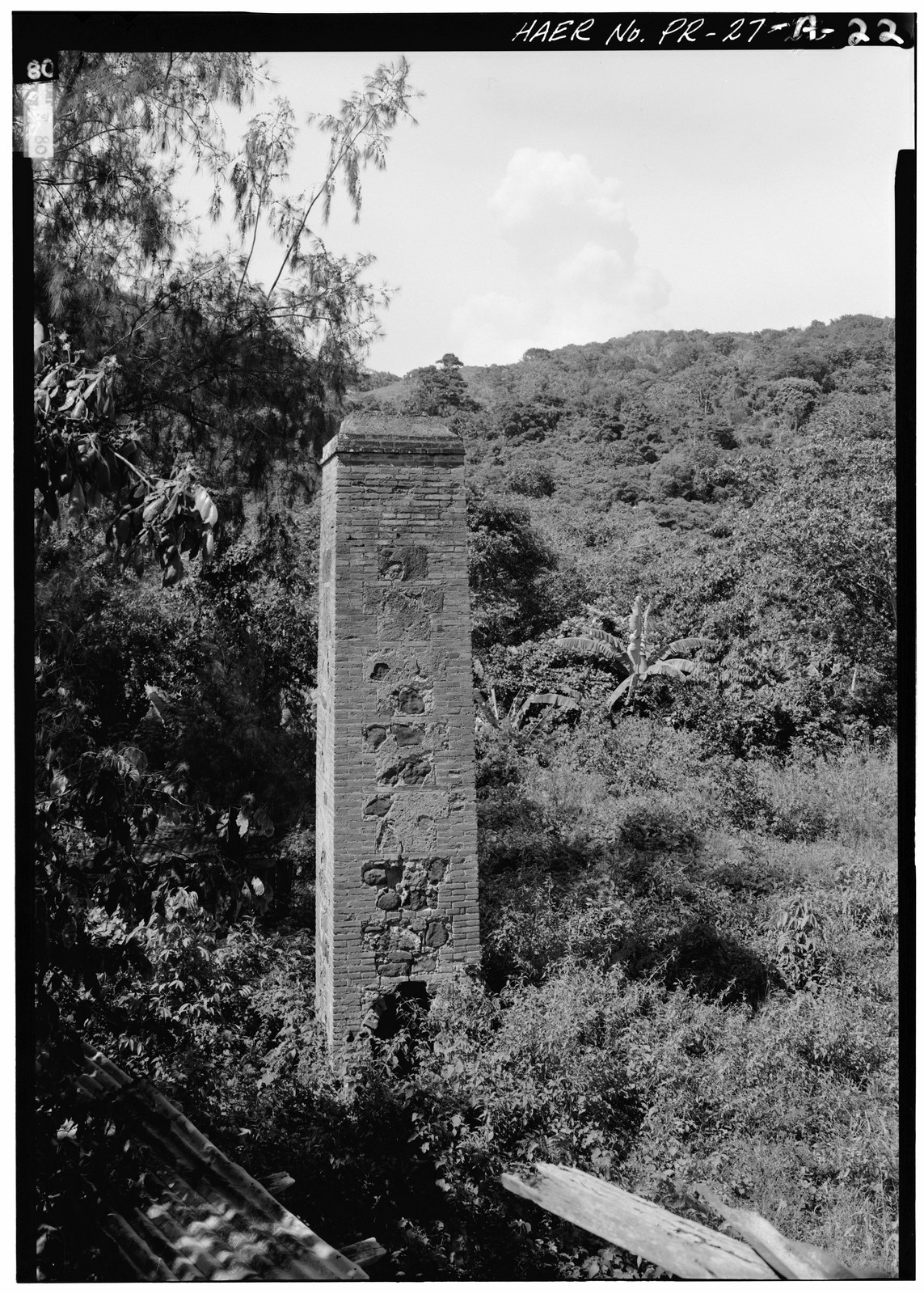 A brick and stone smokestack for a coffee-roasting oven at the Hacienda Santa Clara coffee mill in Yauco, Puerto Rico, standing among trees and banana plants.