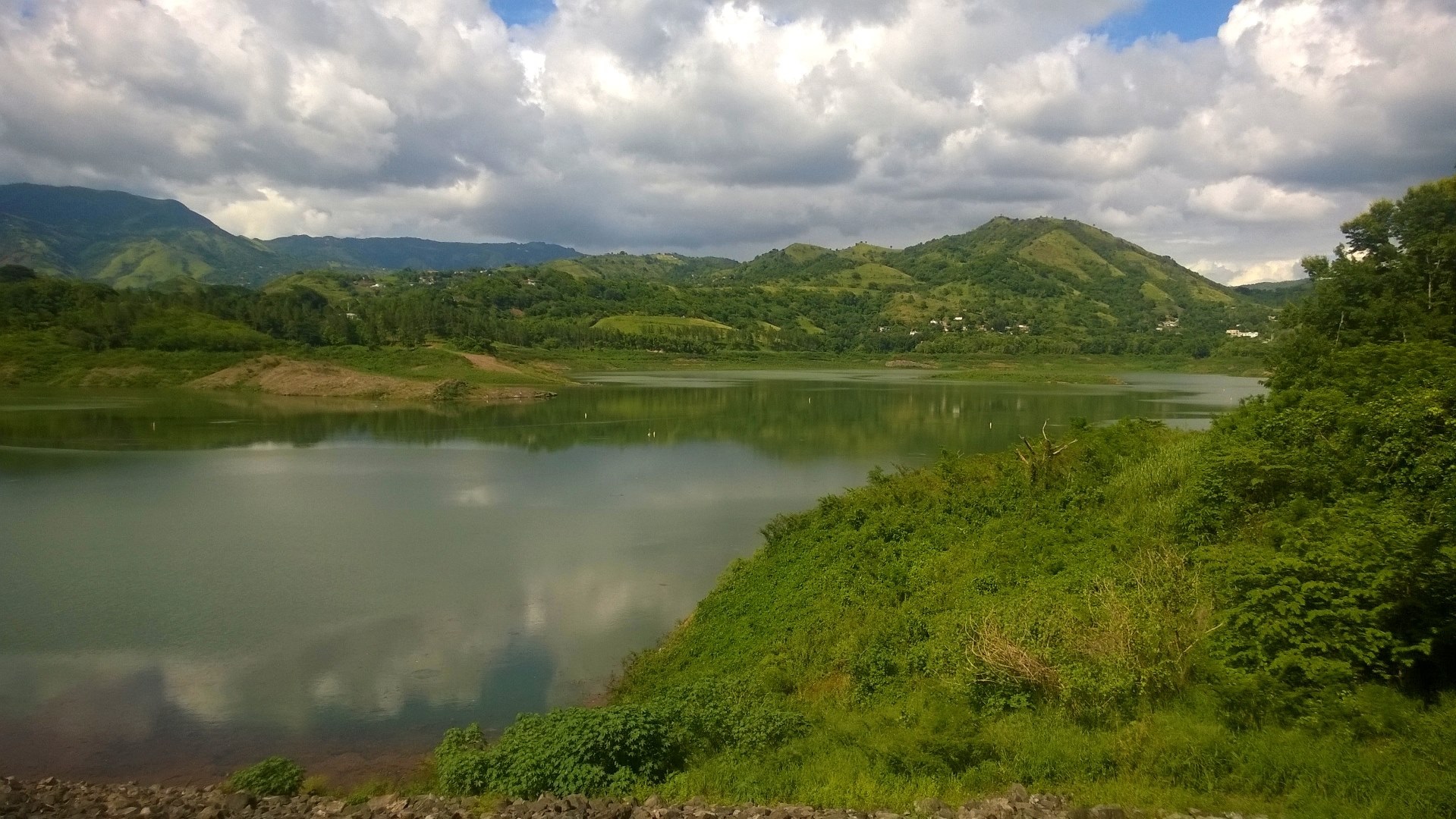 Lago Toa Vaca in Villalba under cumulus cloud, with houses on the green hills across the water and scrub on the near bank.