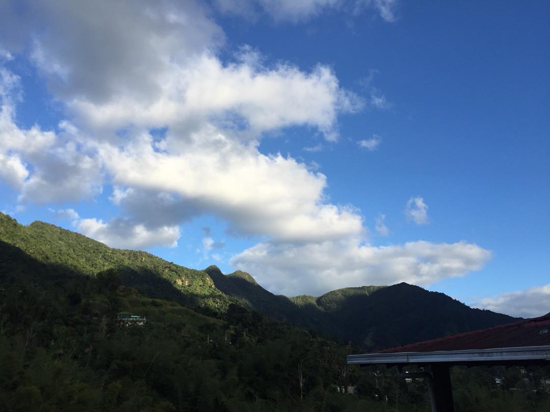 The three peaks of Tres Picachos in Jayuya, Puerto Rico, in afternoon light, with forested slopes below and a tiled roof in the near foreground.