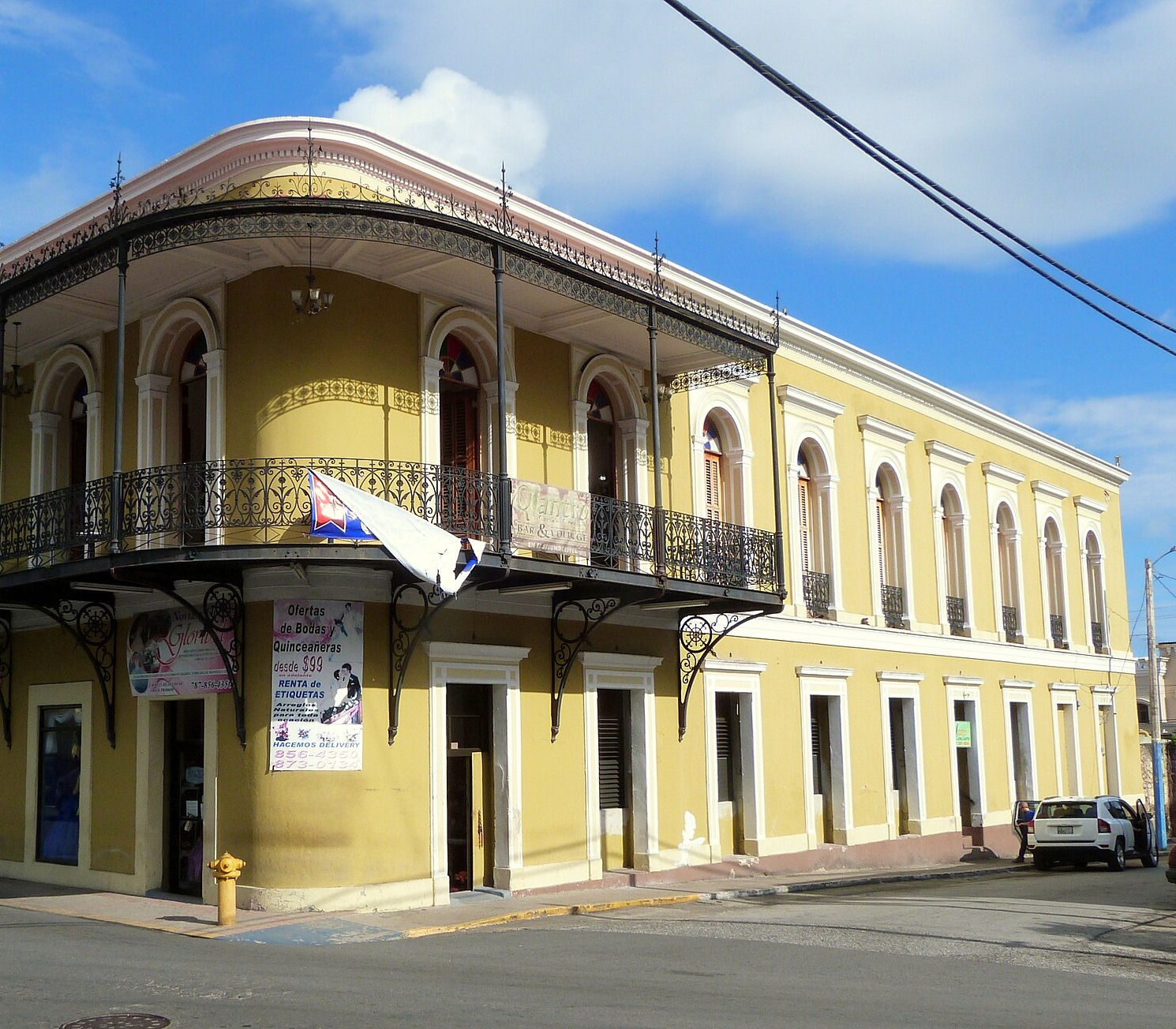 A two-storey yellow corner building with a rounded corner, arched upper windows and a wrought-iron balcony along the upper floor, with shop signs and posters at street level.