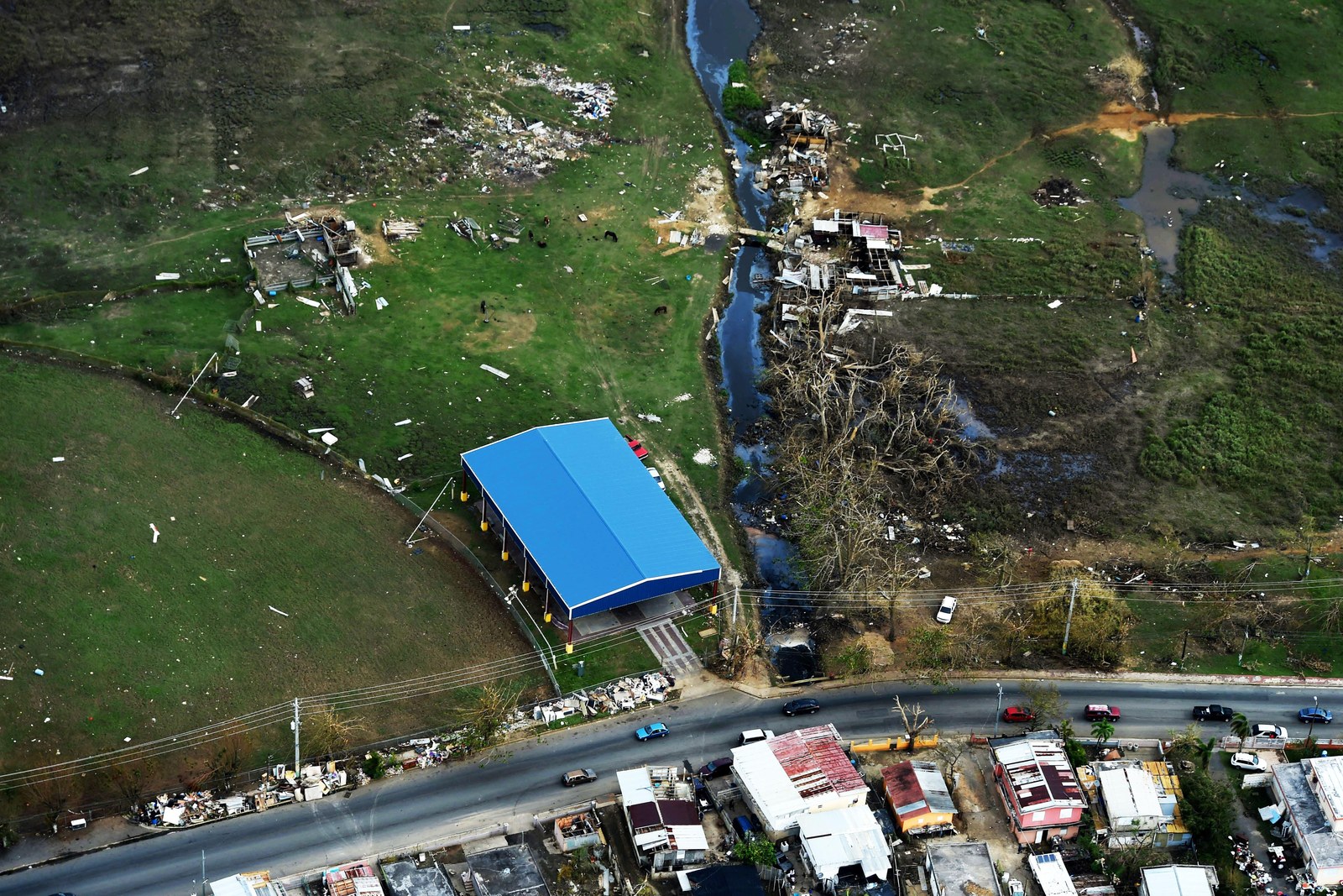 An aerial view of a rural roadside with damaged houses, a stream and debris across green fields.