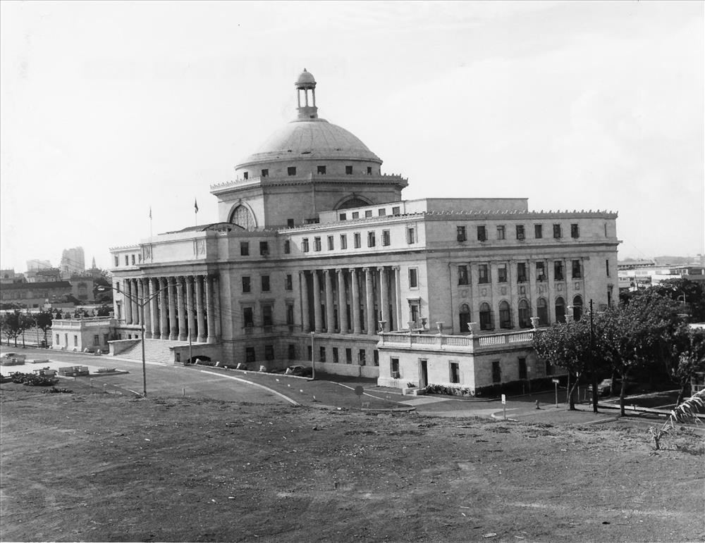 A black-and-white view of the domed Capitol of Puerto Rico across an open lot, with cars in front.