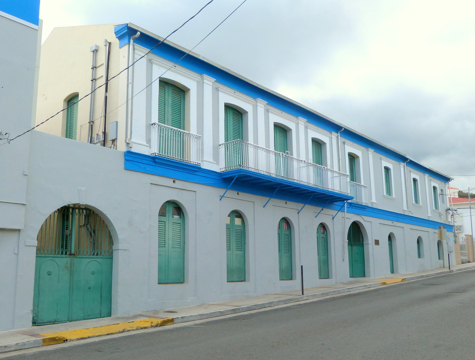 A long two-storey white house with green shutters, an iron balcony and arched doors, on a street.