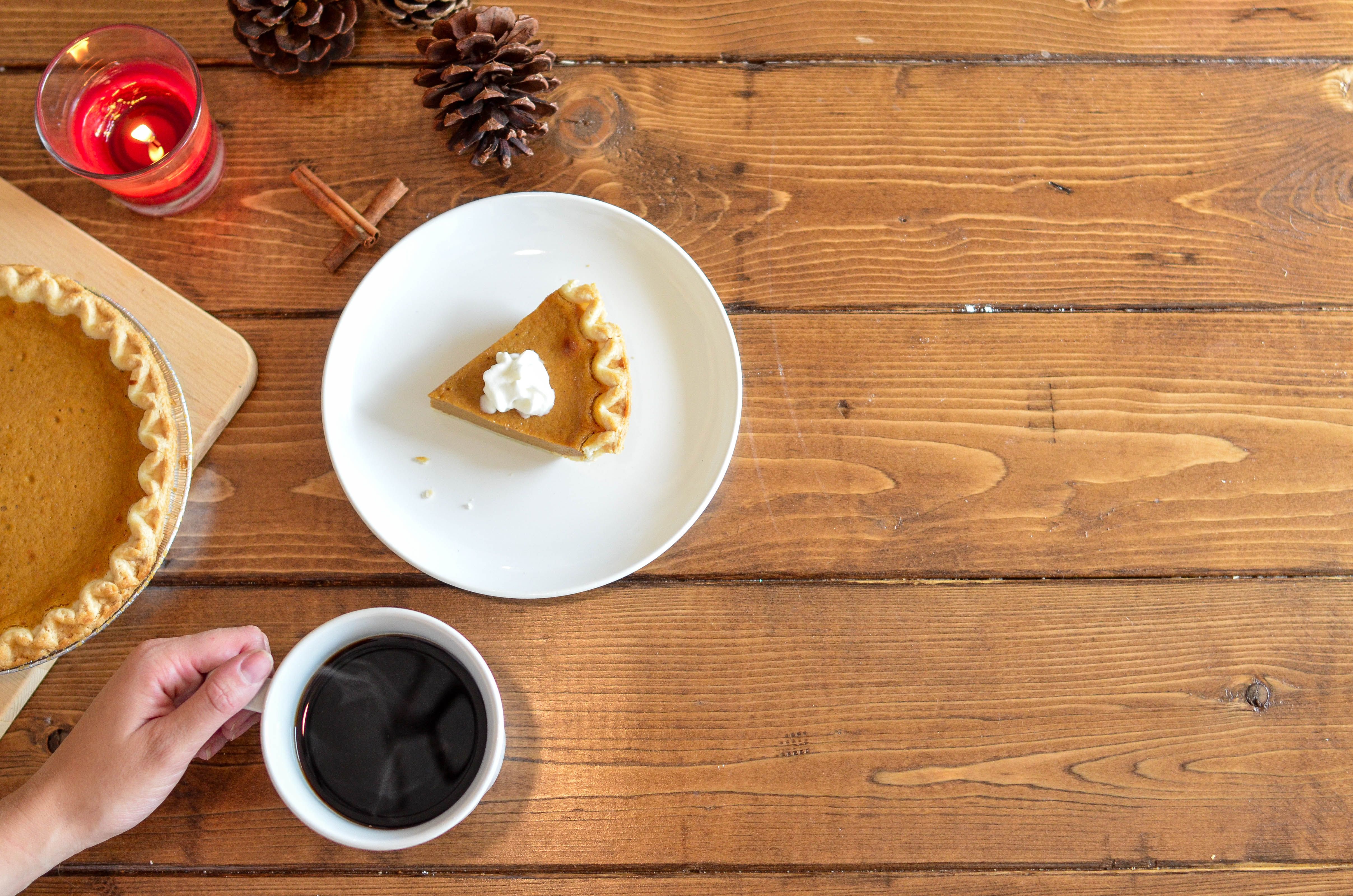 A slice of pumpkin pie with whipped cream and a mug of black coffee on a wooden table with candles and pine cones.