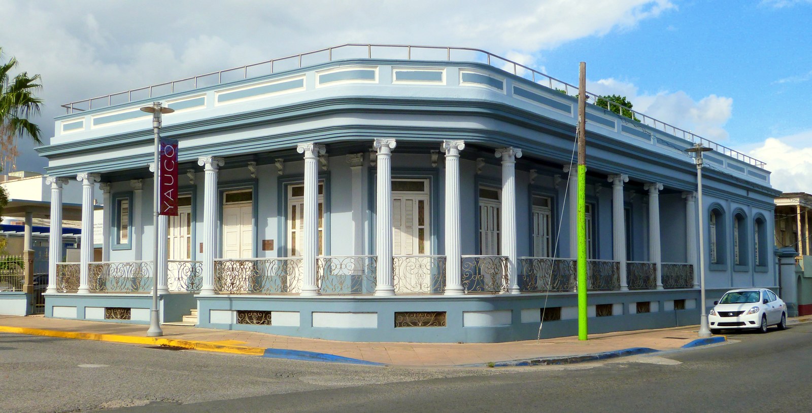 A one-storey corner house with a colonnaded porch and iron railings, beside a street banner reading YAUCO.