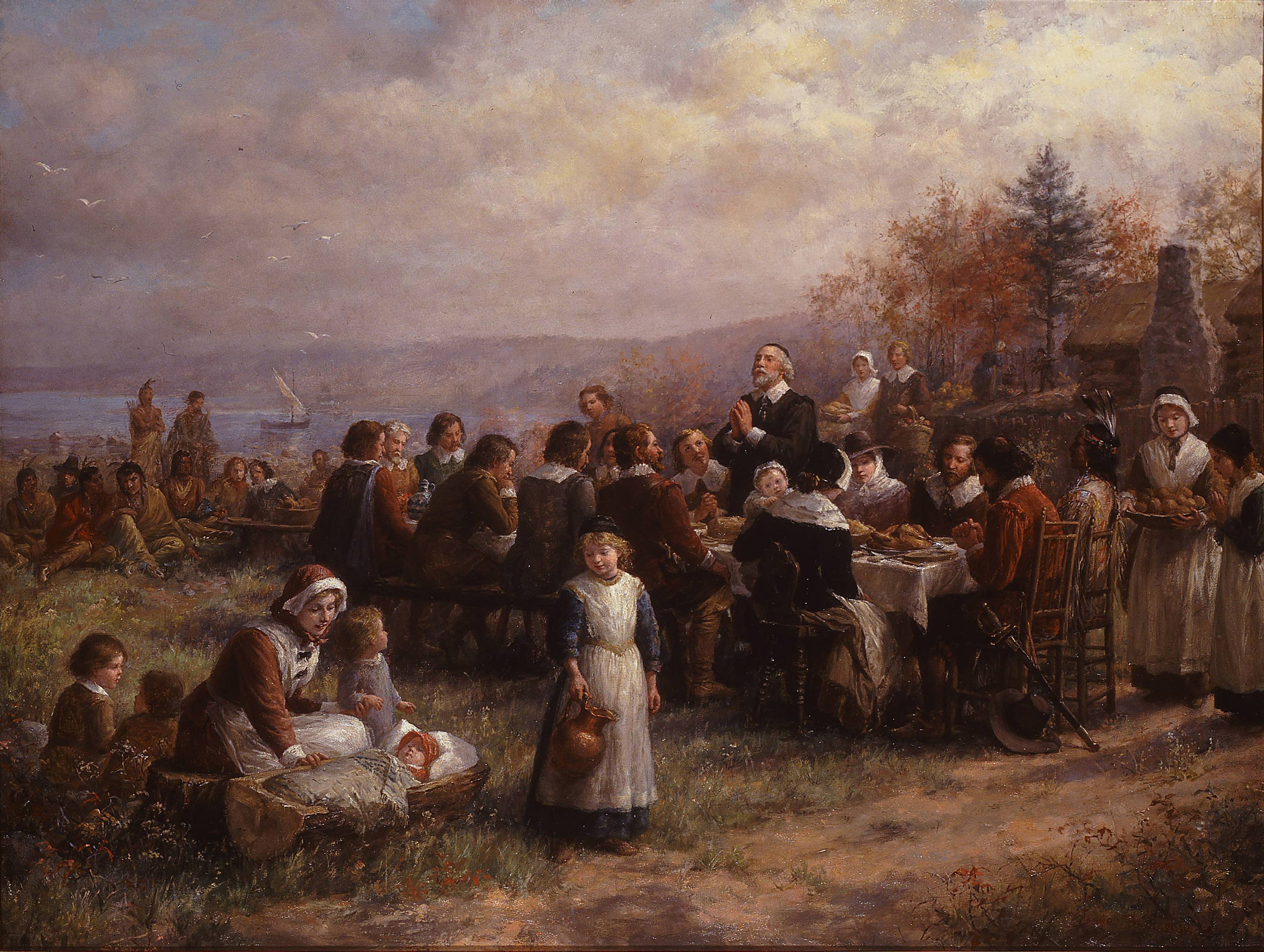 A painting of an outdoor harvest feast at Plymouth, with colonists and Native people around a long table, log cabins and a ship in the bay.